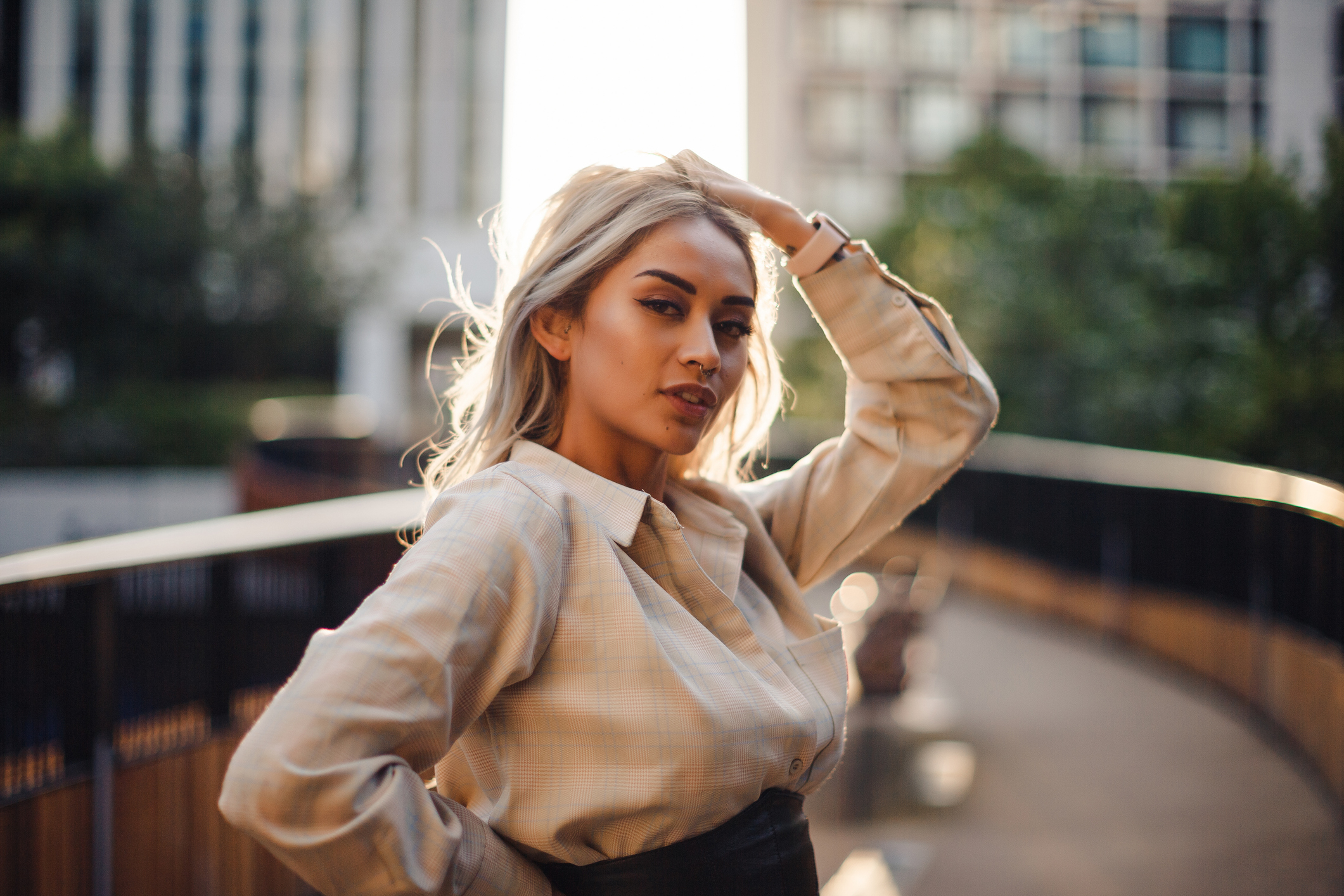 A woman poses confidently on a city sidewalk, with one hand resting on her head. She wears a stylish, long-sleeved blouse. Buildings and trees are in the background