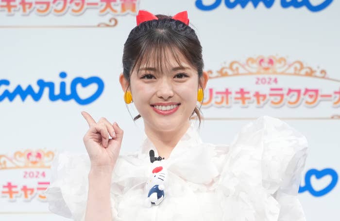 タレントの指原莉乃さんがサンリオキャラクター大賞イベントに参加し、白いドレスを着て手を指し示しています。背景には「Sanrio」と「2024」の文字。