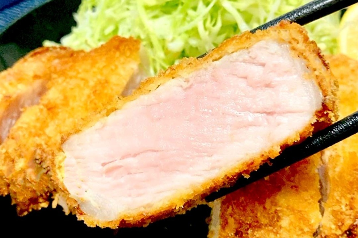 とんかつの断面が見える食事画像。箸で持ち上げた肉の内部はピンク色で、付け合わせに千切りキャベツが盛られています。