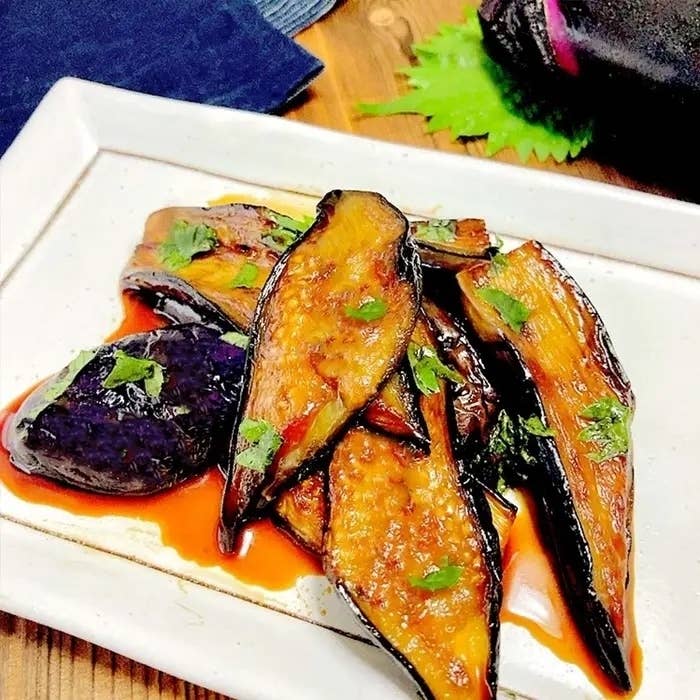 茄子の炒め物が白い皿に盛られている。葉っぱが付け合わせとして置かれ、香草が散らされている。