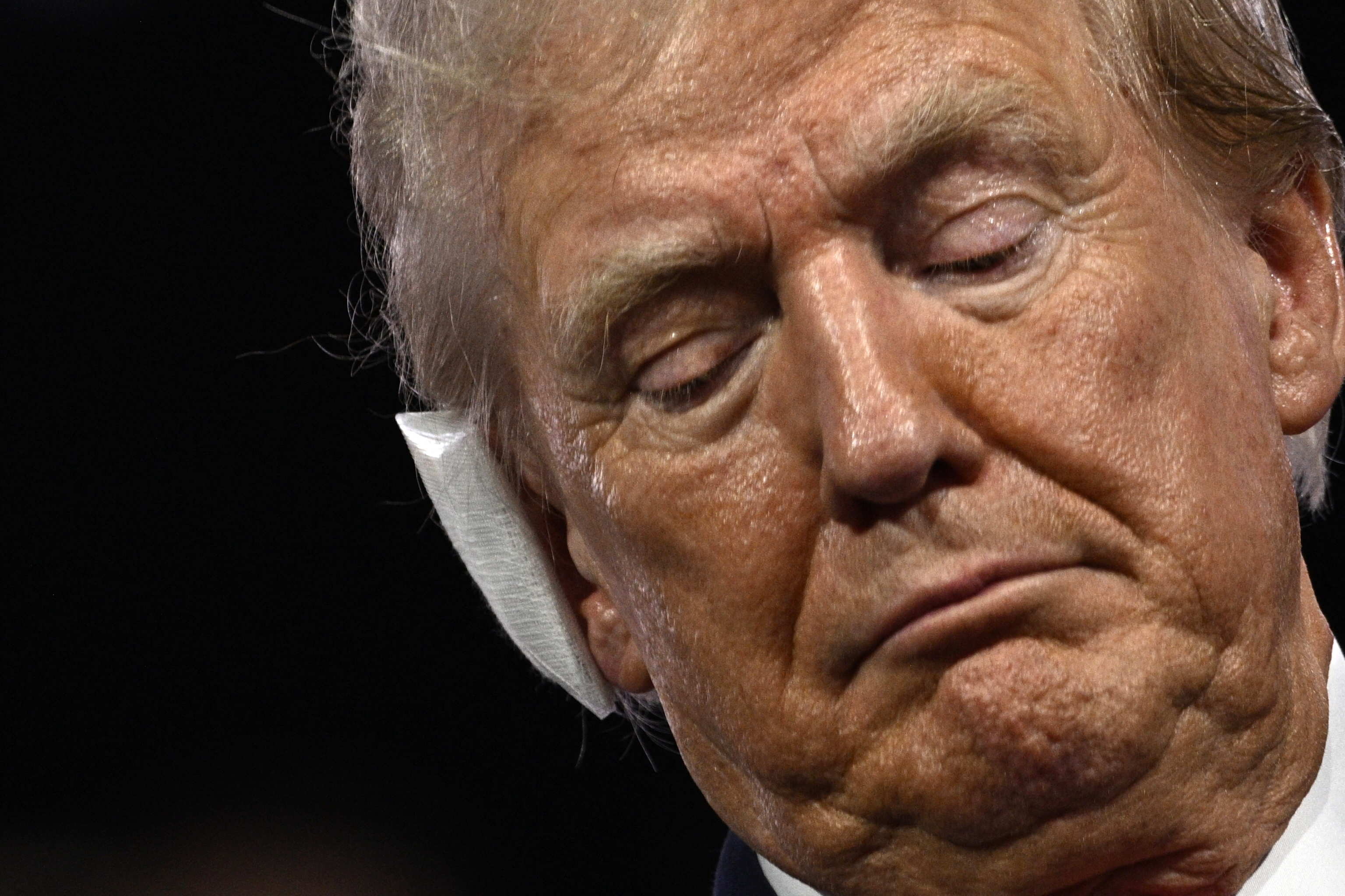Donald Trump with his eyes closed, looking downward, a piece of tissue or paper stuck to the side of his head