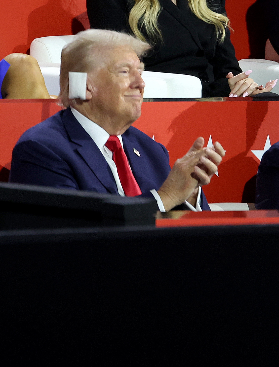 Donald Trump sits clapping and smiling, wearing a suit with a red tie. Visible is a piece of what appears to be tissue or similar material on the side of his head