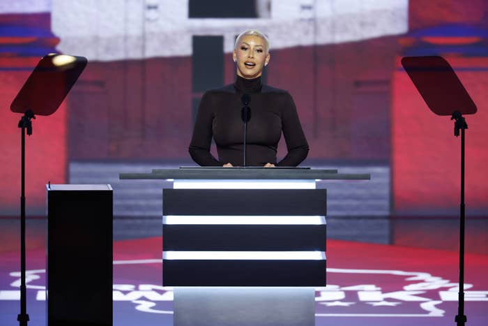 A woman stands at a podium, speaking into a microphone, with teleprompters on either side. She wears a form-fitting, long-sleeve dress