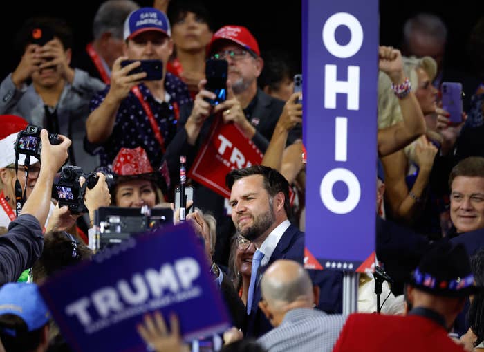 J.D. Vance standing in a crowd of supporters, surrounded by people holding "TRUMP" signs and taking photos, with an "OHIO" sign in the background