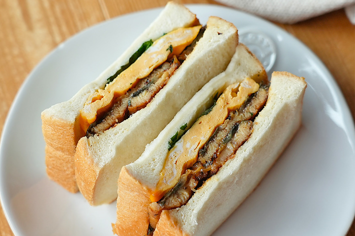 白い皿に盛られた2つのサンドイッチ。卵、肉、野菜が厚めのパンに挟まれている。背景には布が見える。