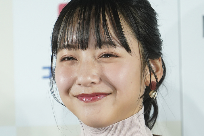 女性が微笑んでカメラを見つめている。彼女はタートルネックのニットとチェック柄のベストを着用している。