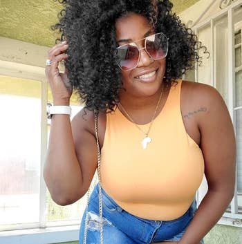 reviewer wearing an orangish sleeveless bodysuit and jeans, accessorized with sunglasses and a necklace