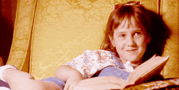A young girl, Matilda Wormwood, is lying on a couch and smiling while holding an open book. She is wearing a floral shirt and overalls with a bow in her hair