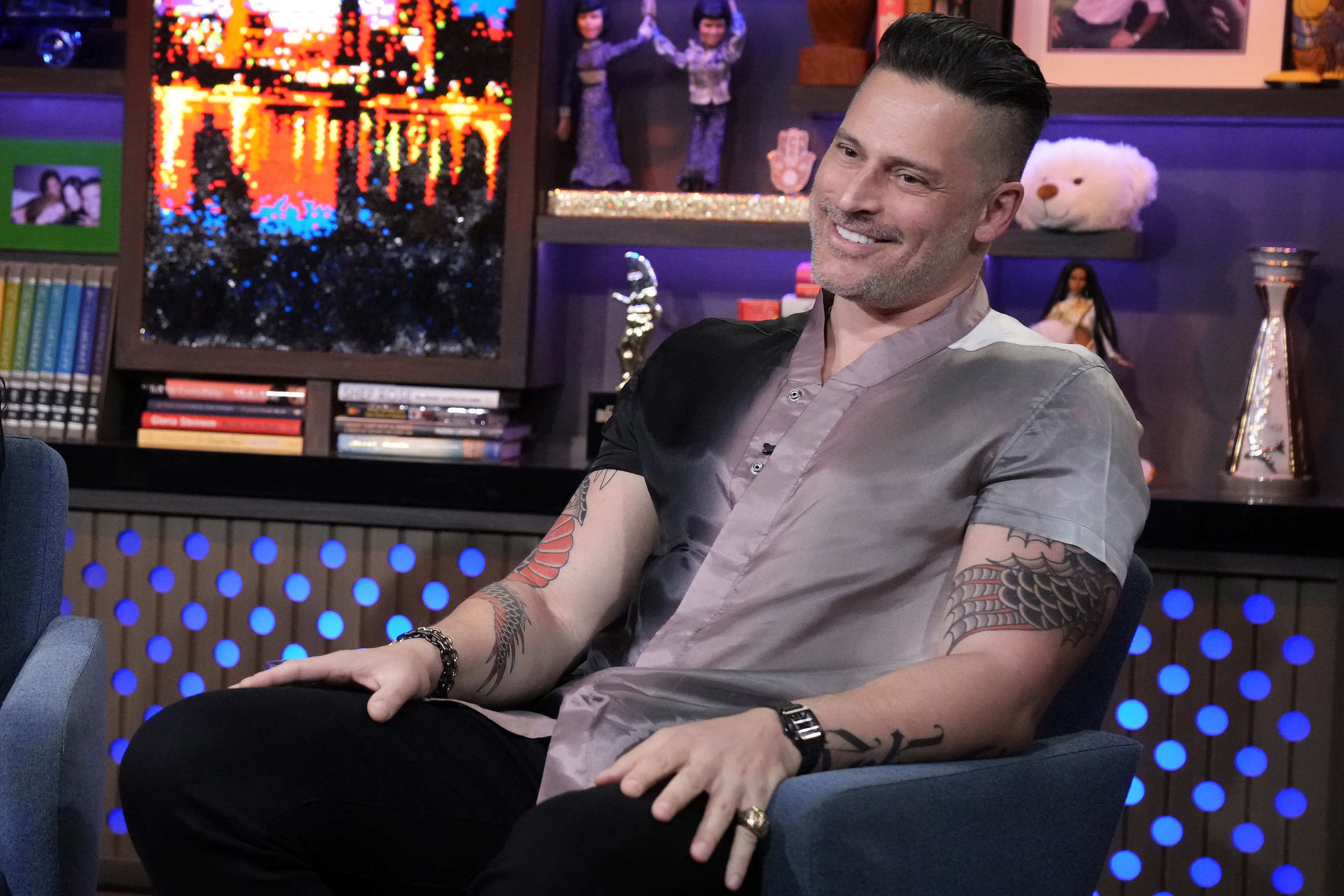 Joe Manganiello is seated on a talk show set, wearing a casual shirt. There is a colorful cityscape painting and various decor in the background