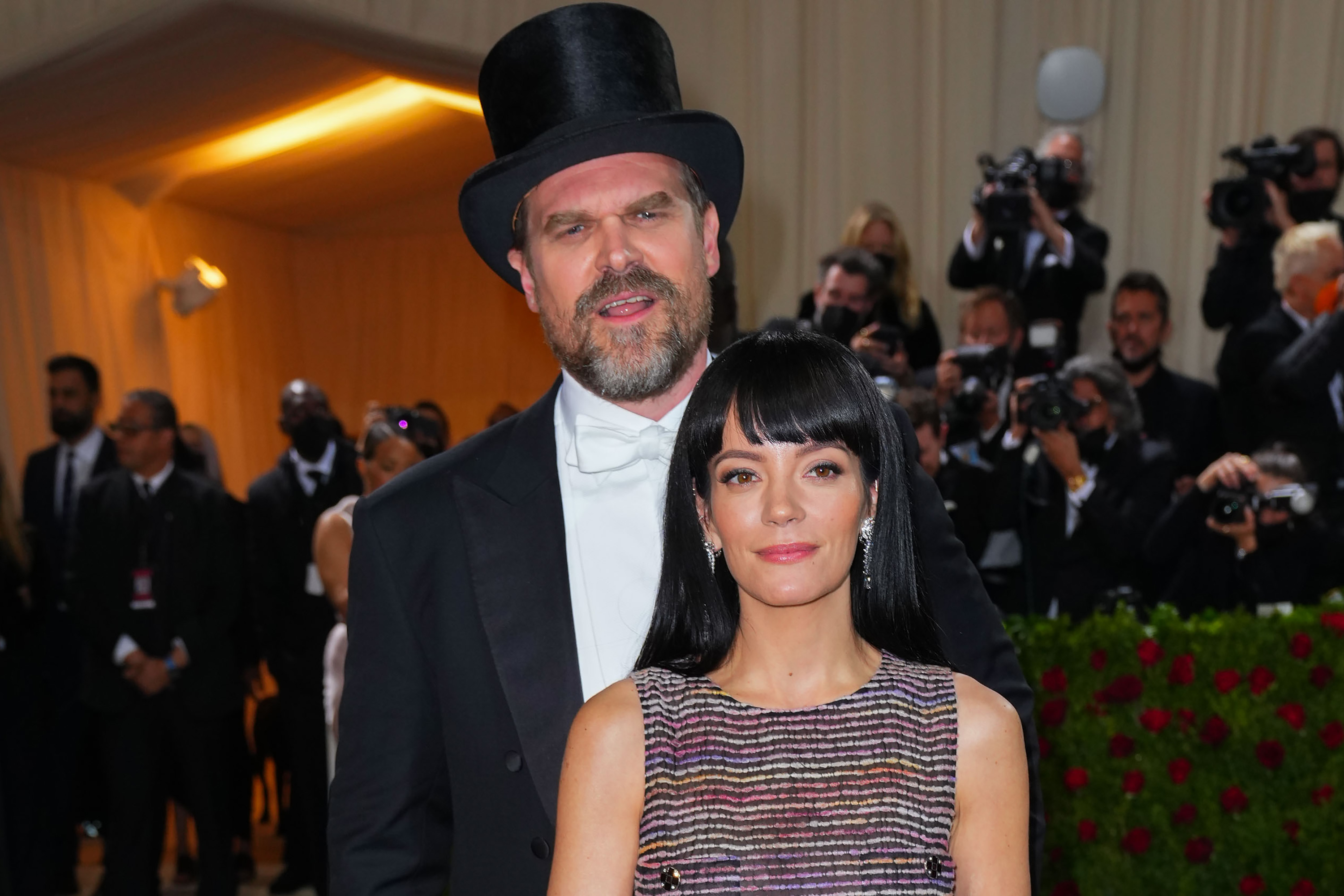 David Harbour in a top hat and formal wear with Lily Allen in a sleeveless, glittery gown at a formal event with photographers in the background