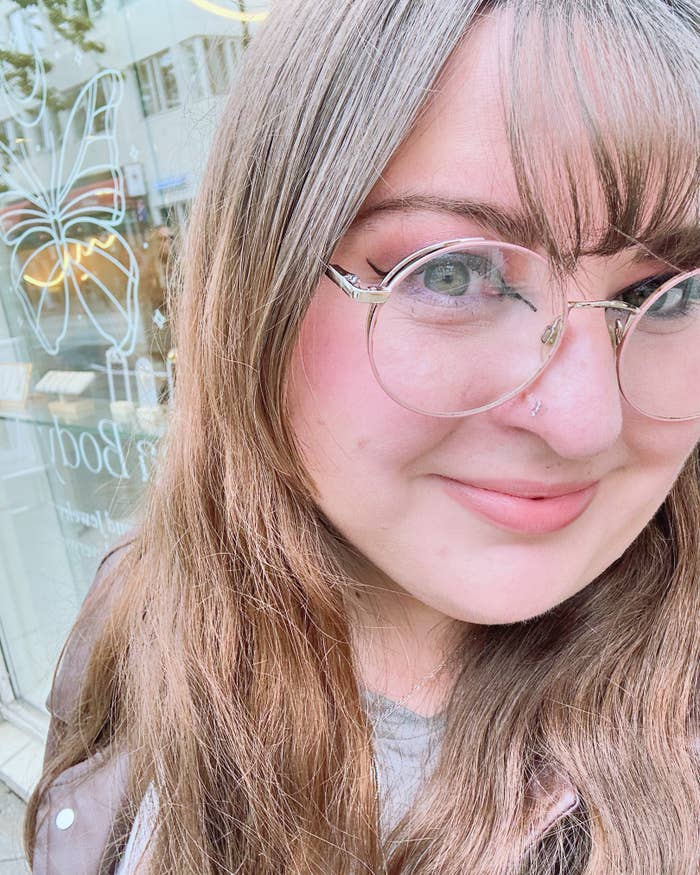 A woman with long hair, glasses, and a nose piercing smiles for a close-up selfie in front of a shop window with a butterfly design