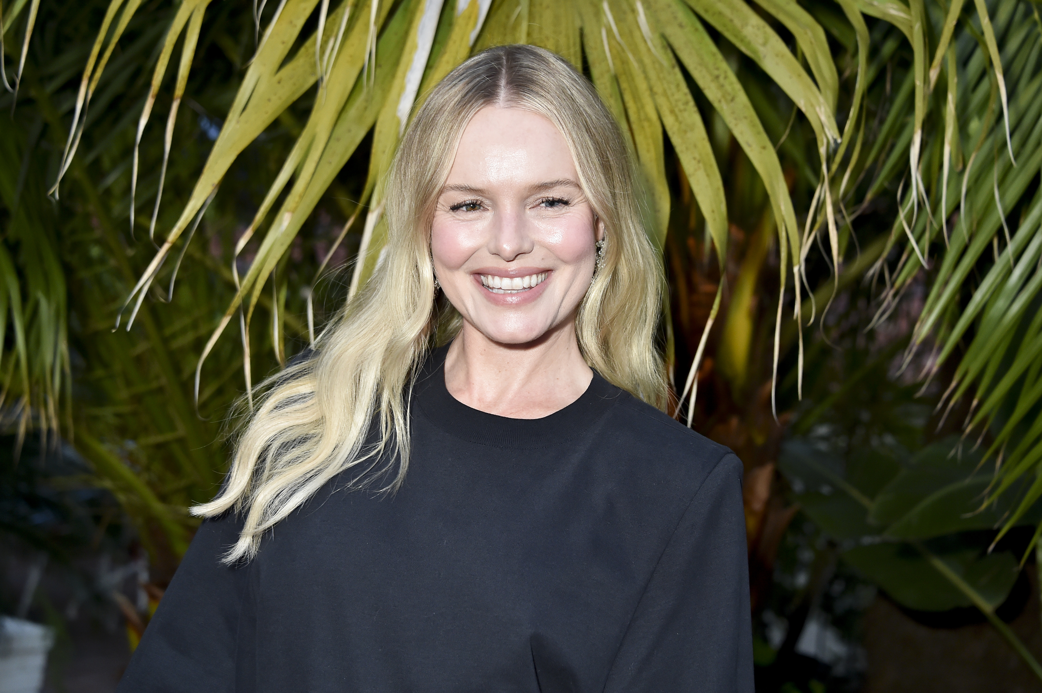 Kate Bosworth with long blonde hair, wearing a simple, elegant outfit, smiles in front of lush green palm leaves