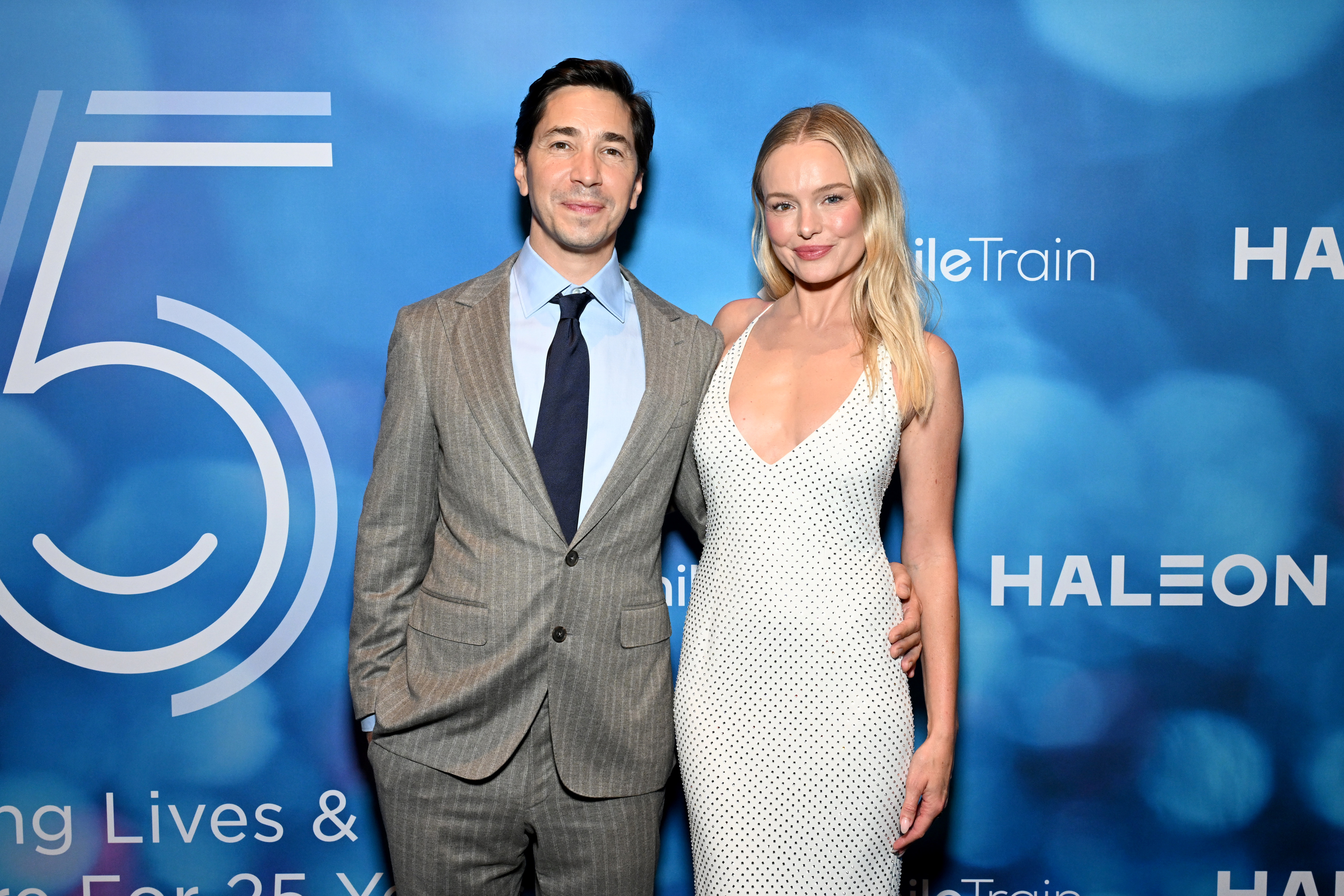 Justin Long in a suit and Kate Bosworth in a sleeveless, fitted dress pose together at an event in front of a blue backdrop