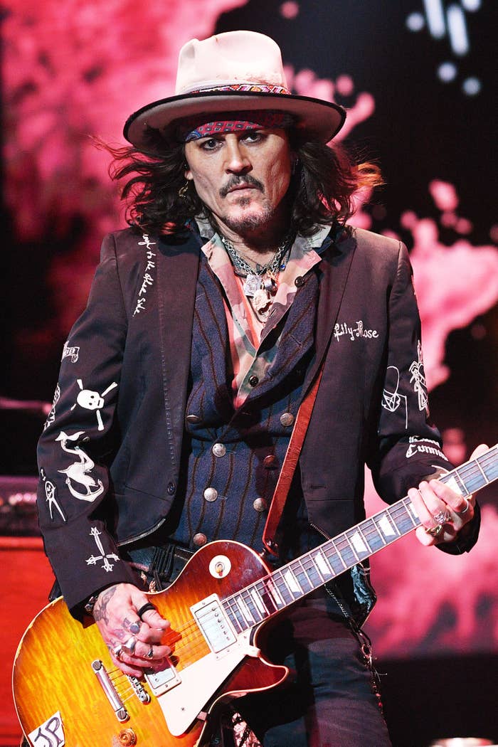 Johnny Depp playing guitar, wearing a hat and an embroidered jacket, on stage