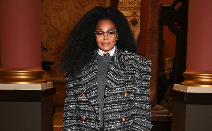 Janet Jackson stands indoors wearing a long, patterned coat over a gray sweater and shirt, holding a black handbag