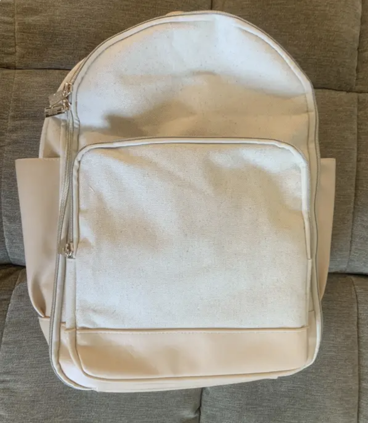 A light-colored travel backpack with a front pocket and multiple zippers, placed on a cushioned surface
