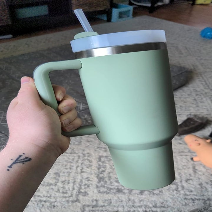 Person holding a large, mint green insulated tumbler with a straw and handle. In the background, a toy pig lies on the floor