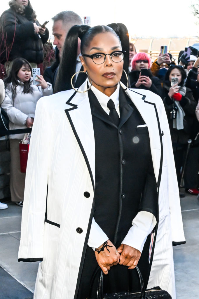 Janet Jackson in a pinstripe blazer over an outfit with a tie, hoop earrings, and ponytails, poses in front of a crowd
