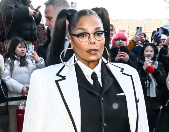 Janet Jackson in a pinstripe blazer over an outfit with a tie, hoop earrings, and ponytails, poses in front of a crowd
