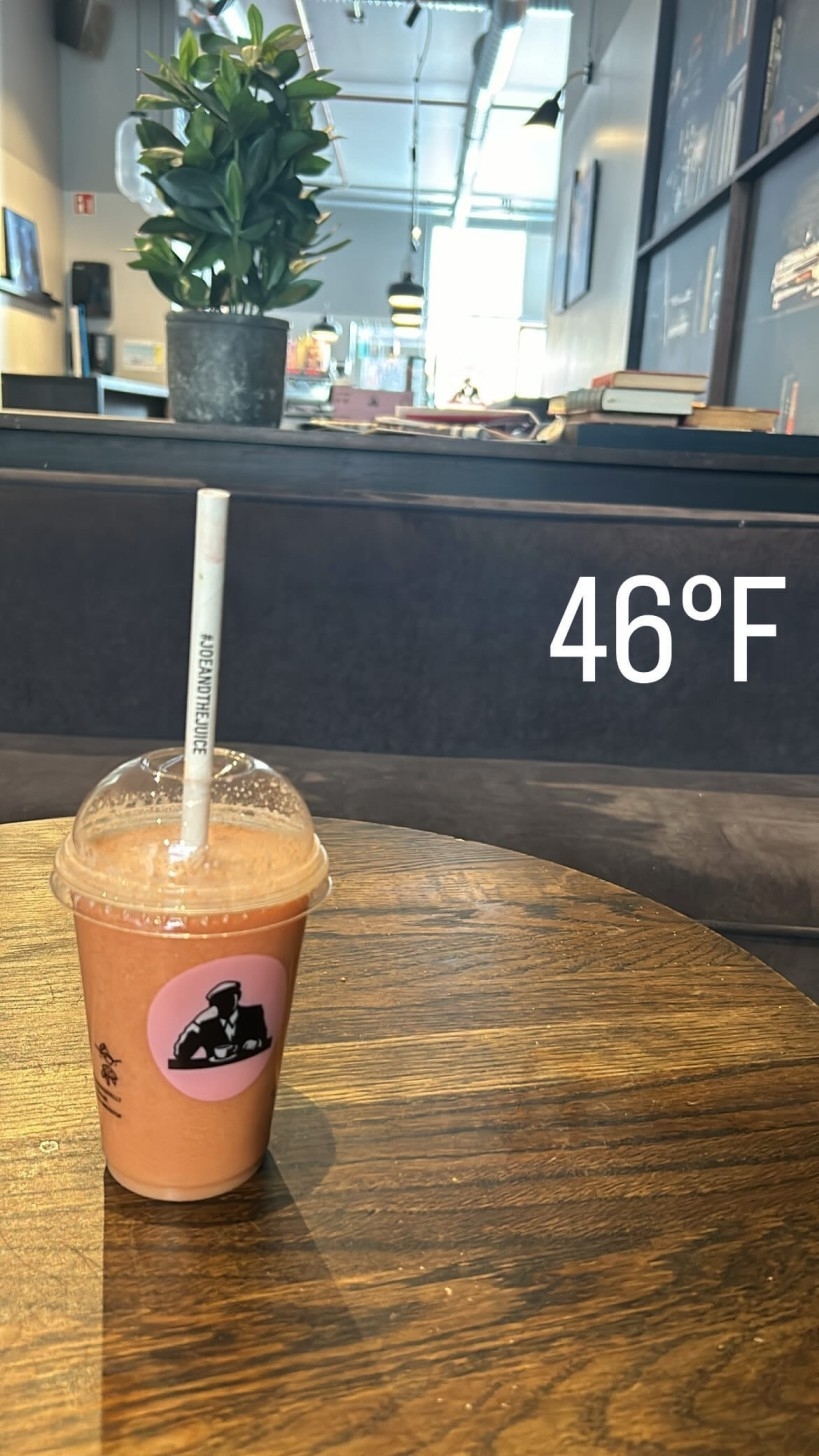 A plastic-cupped smoothie with a straw sits on a wooden table in a modern cafe. A plant is in the background. The temperature reads 46°F