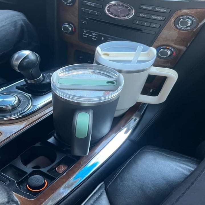 Two large reusable travel cups, one black and one white, with handles and lids, are placed in the cup holders of a car's center console
