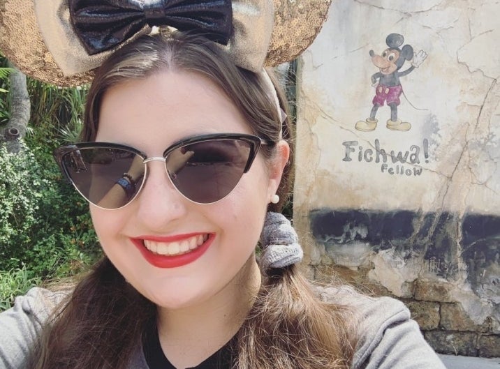 Kristen wearing sunglasses and Minnie Mouse ears smiles, standing in front of a mural of Mickey Mouse at a Disney theme park