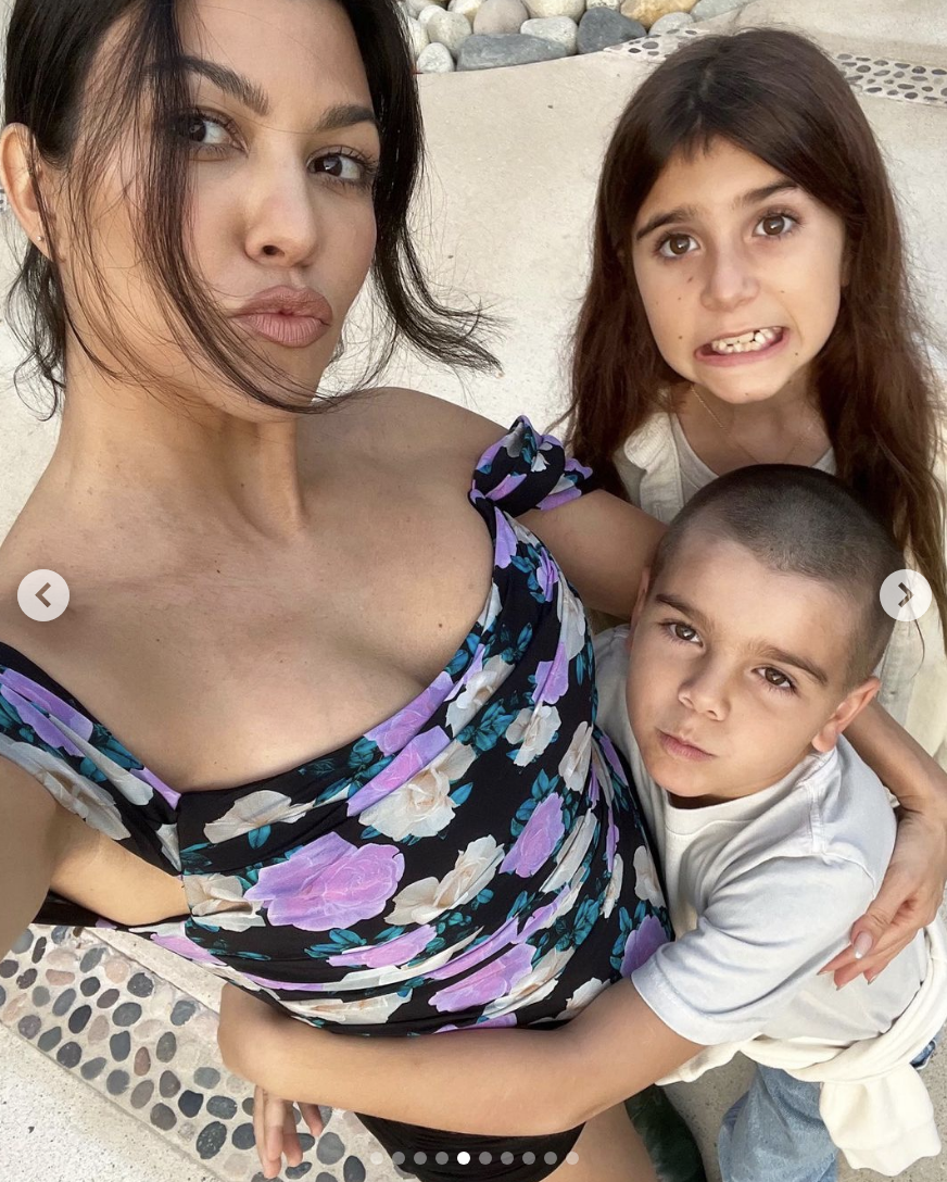 Kourtney Kardashian in a floral dress, taking a selfie with her children, Penelope and Reign, who are hugging her