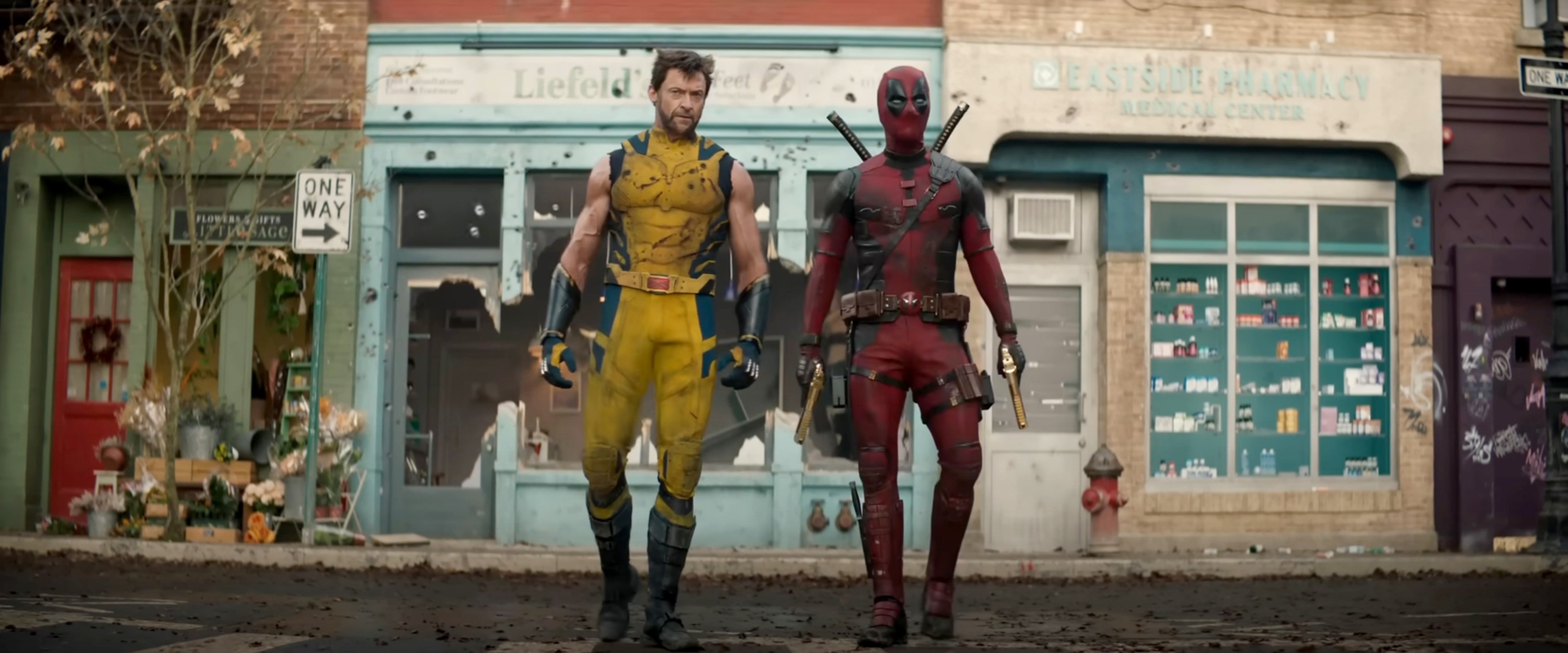 Hugh Jackman as Wolverine in a yellow suit and Ryan Reynolds as Deadpool in a red suit are walking side by side on a street with shops in the background