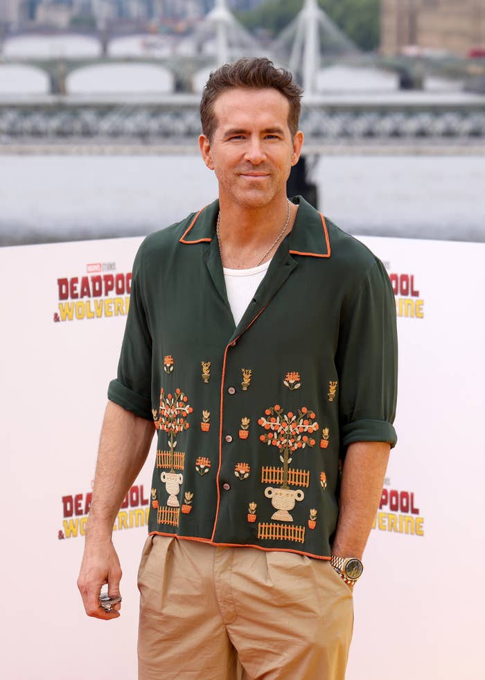 Ryan Reynolds at a press event, wearing a casual shirt with a nature-themed design and beige pants, promoting "Deadpool & Wolverine"