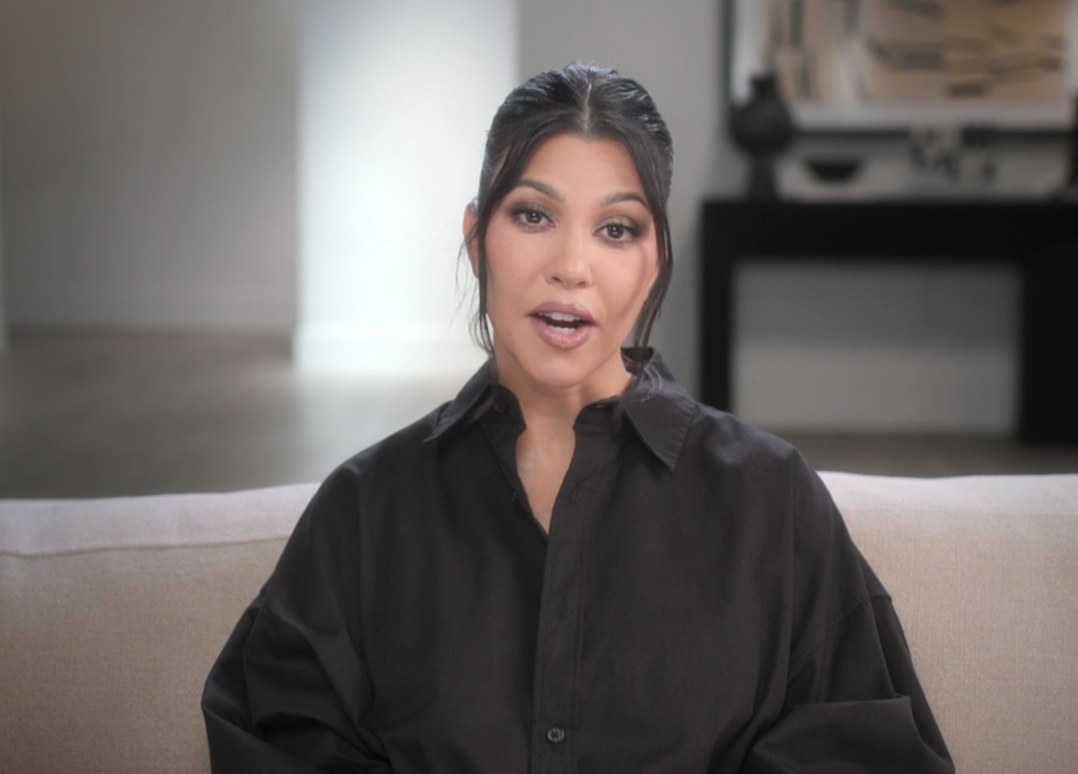 Kourtney Kardashian is sitting on a beige couch in a living room, wearing a black shirt with her hair tied back. She appears to be speaking