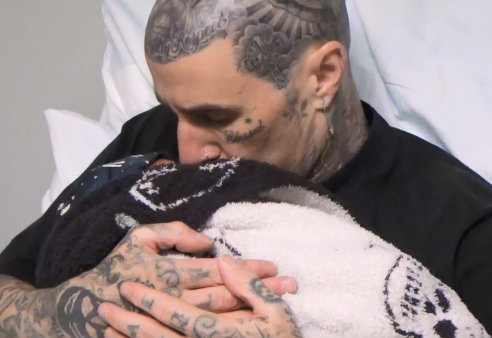 Travis Barker, with extensive tattoos, kisses his newborn baby while holding them close, wrapped in a patterned blanket