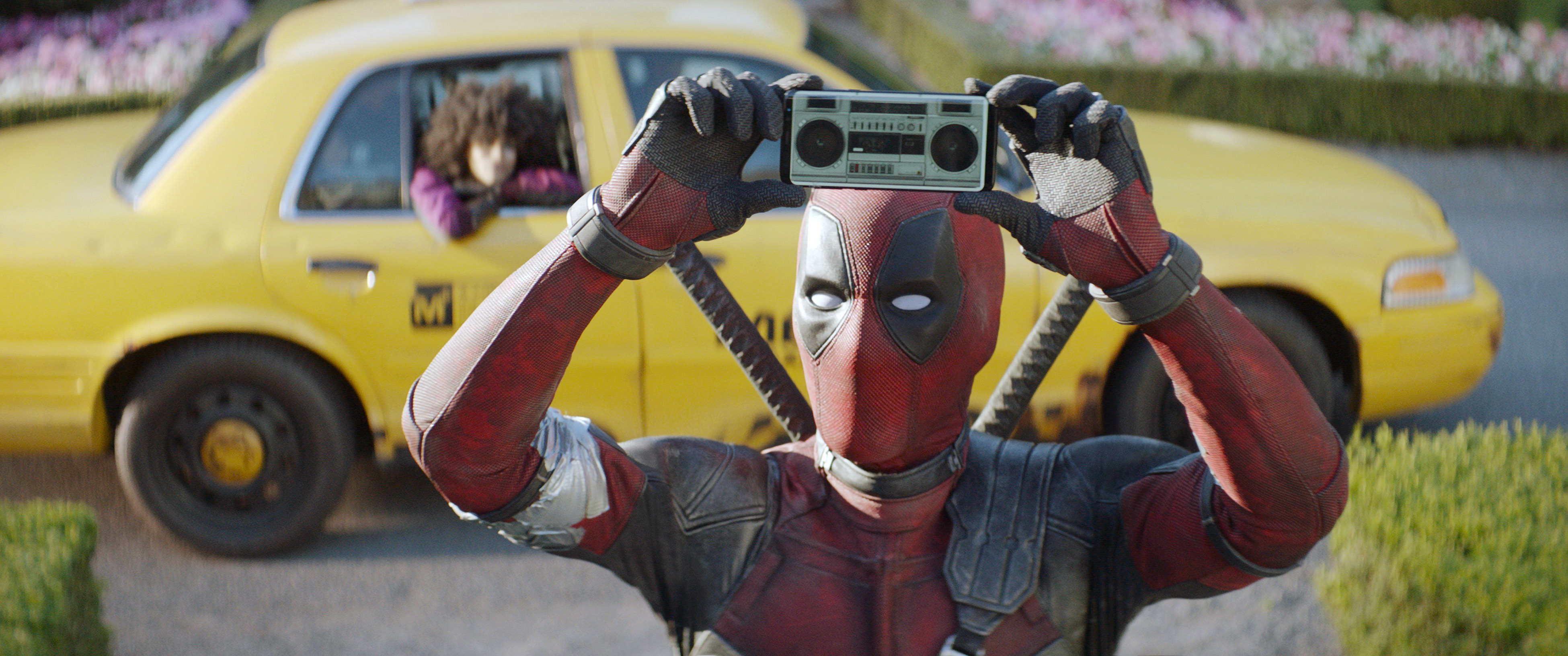 Deadpool holds a boombox over his head in front of a taxi with Negasonic Teenage Warhead looking out the window