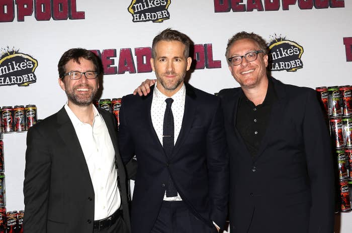 Rob Liefeld, Ryan Reynolds, and Rhett Reese at a Deadpool event. Ryan wears a dark suit, white shirt, and tie. Rob and Rhett are in dark suits with no ties