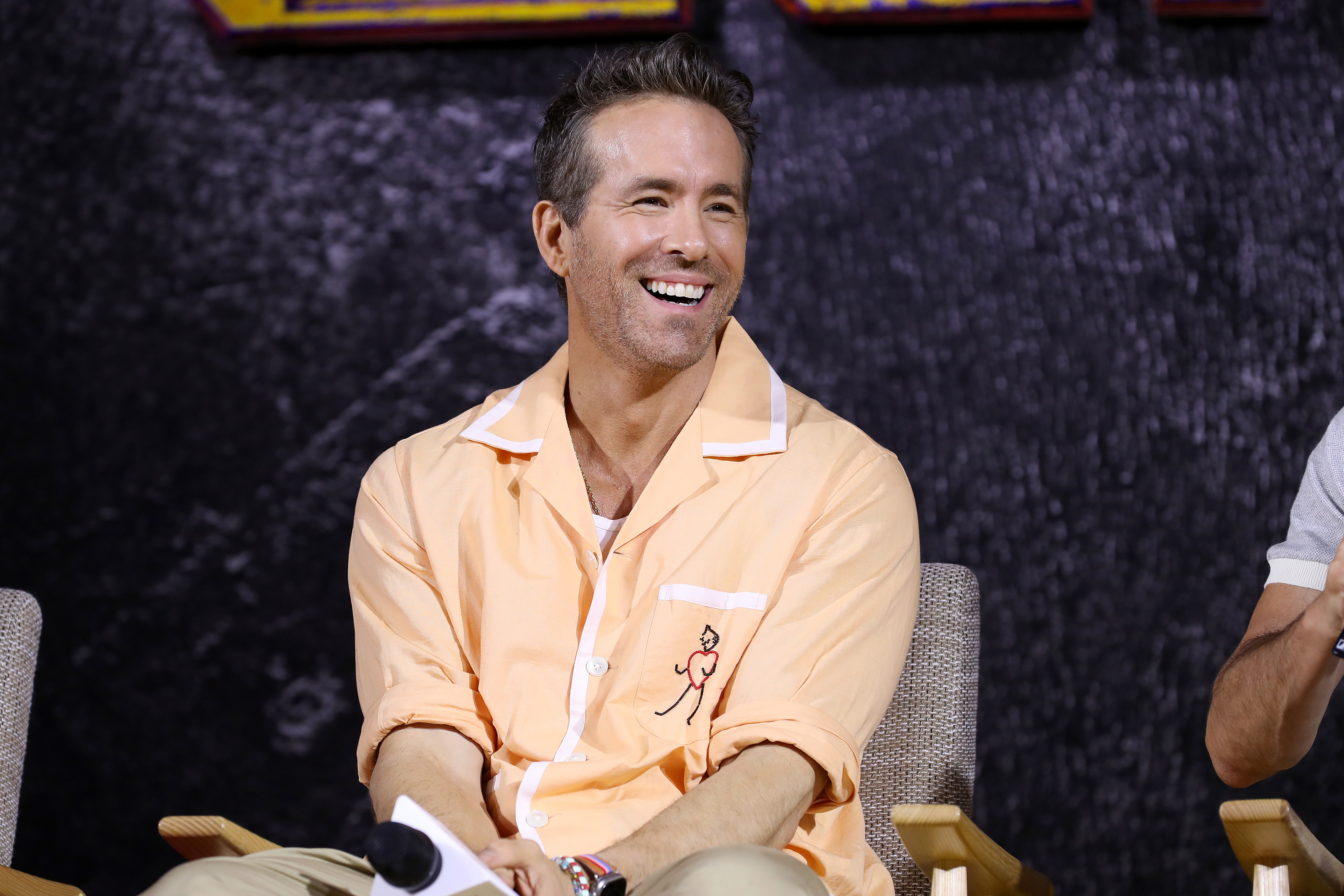 Ryan Reynolds sits and smiles, wearing a casual shirt and holding a microphone
