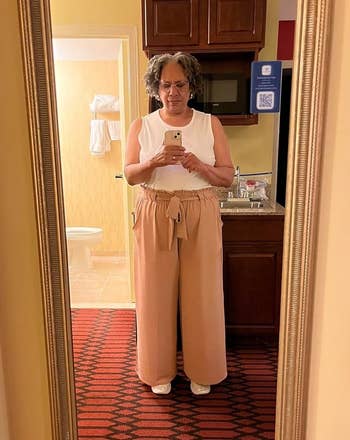 A reviewer takes a full-length reflector selfie successful a bathroom. She wears a achromatic sleeveless top, high-waisted wide-leg pants with a belt, and achromatic shoes