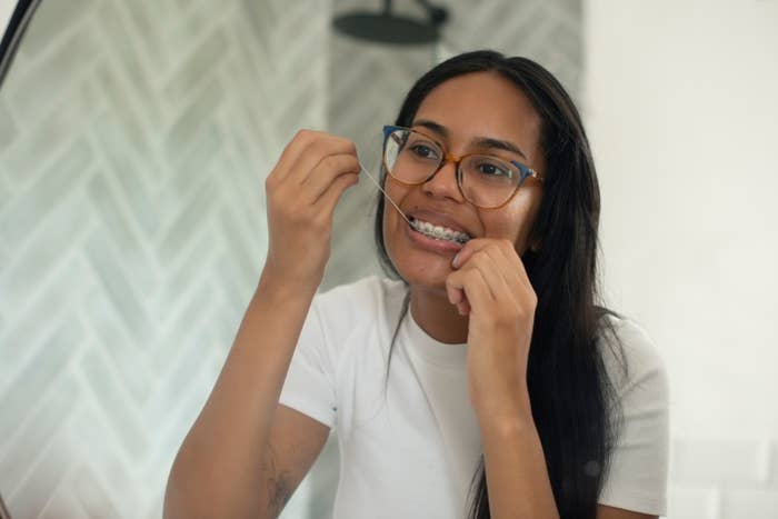 A person with braces and glasses, wearing a white shirt, smiling and flossing their teeth in front of a mirror