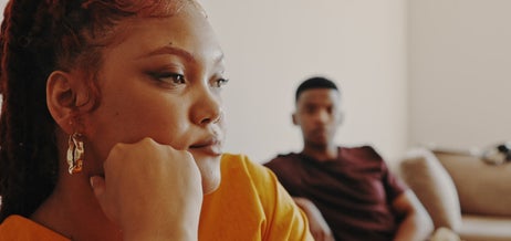 A woman with braids and wearing a stylish outfit looks thoughtful while sitting on a couch; a man in casual clothes sits on another couch in the background