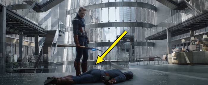 Captain America holding a hammer, looking down at a fallen Captain America in a modern, high-tech building with glass and metal structures