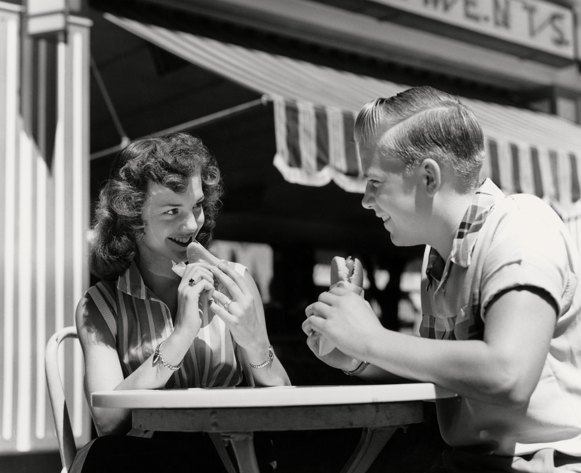 A couple in a vintage diner gazes at each other lovingly while sharing sandwiches. Both are seated at a small table outdoors