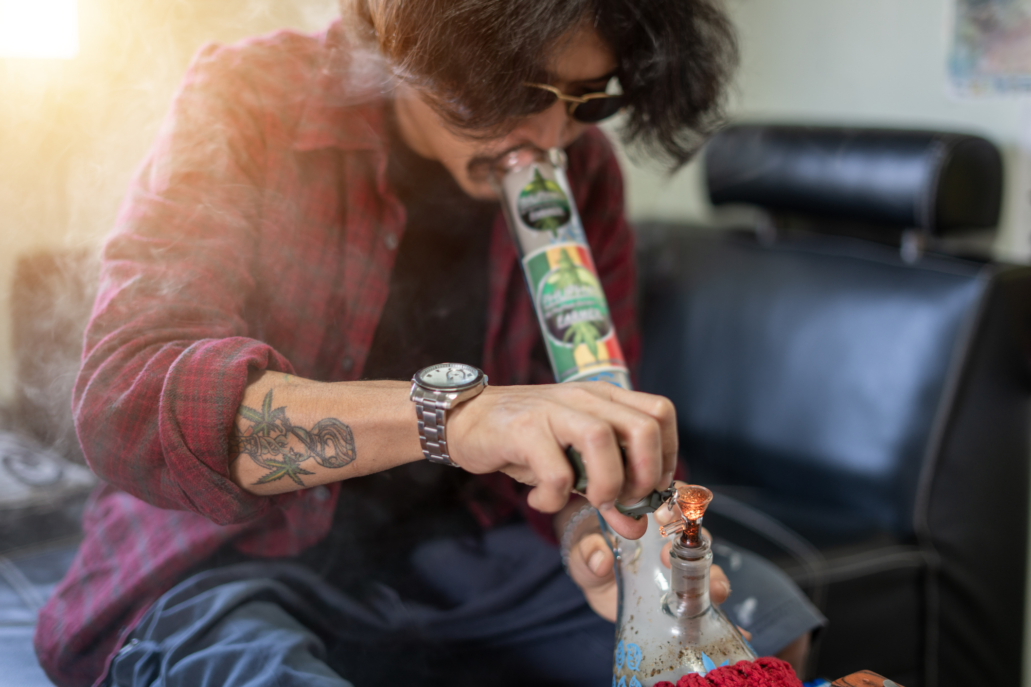 A person with sunglasses and a tattoo, wearing a plaid shirt and wristwatch, is smoking from a bong while seated indoors