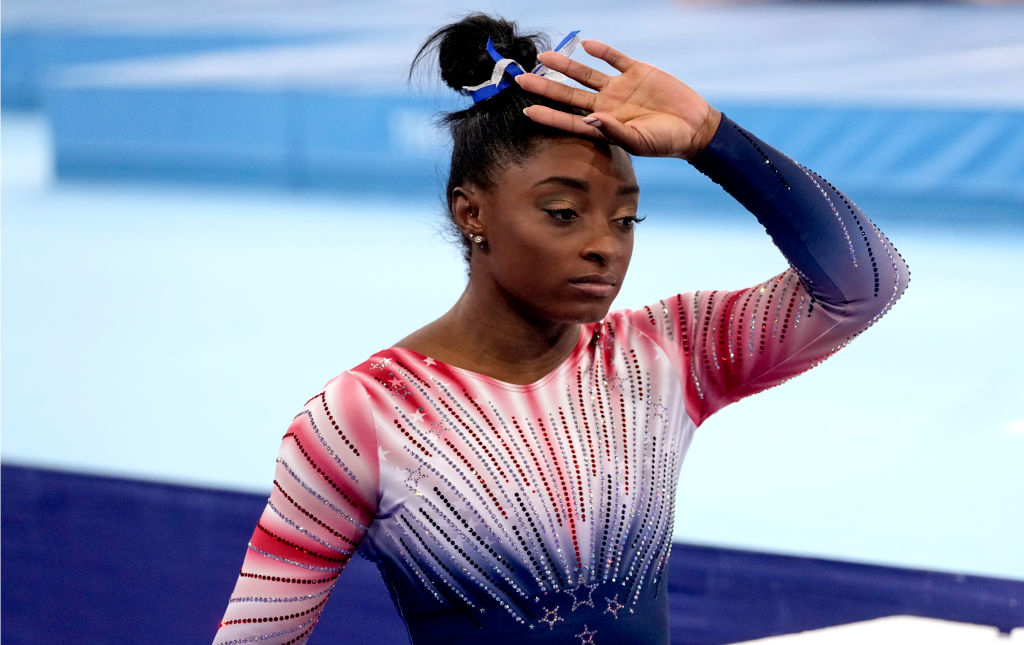 Simone Biles in a gymnastics leotard, raising her hand to her forehead, appears focused during a performance or competition