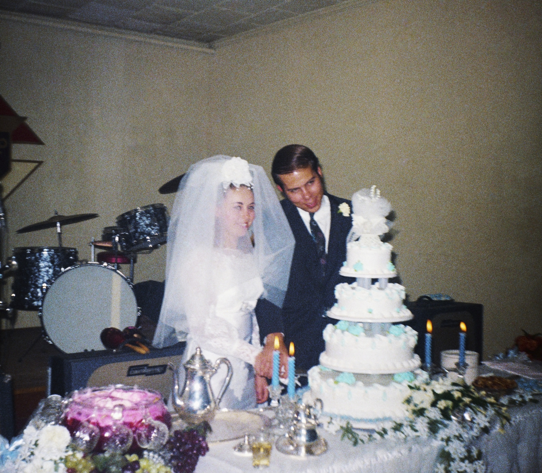 The image shows a couple, with the woman in a wedding dress and veil and the man in a suit, cutting a multi-tiered wedding cake at their wedding reception