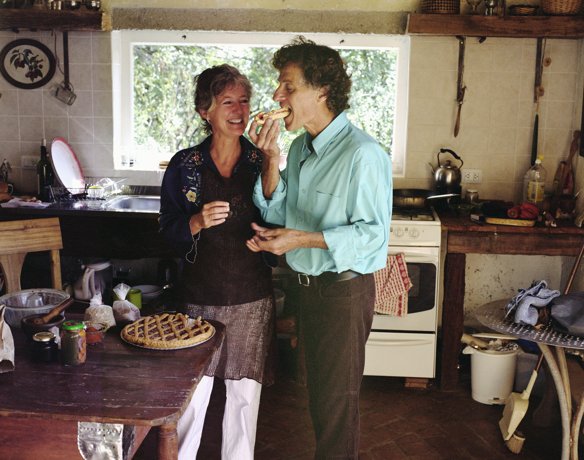 An older couple, with the man wearing a blue shirt, share a slice of pie in a rustic kitchen, smiling and enjoying each other's company