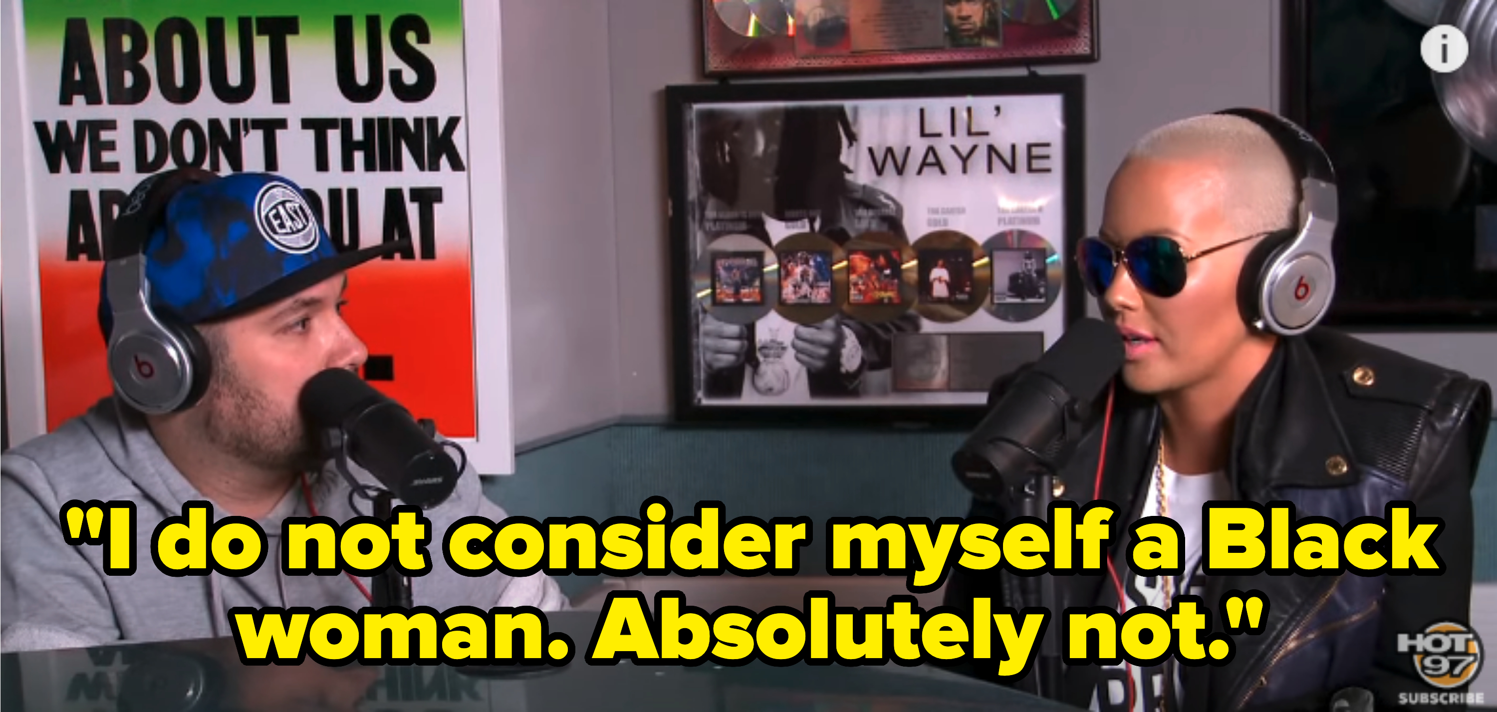 Amber Rose, wearing sunglasses and a leather jacket, is interviewed by a man in a hoodie and cap on HOT 97 radio station. Posters are visible in the background