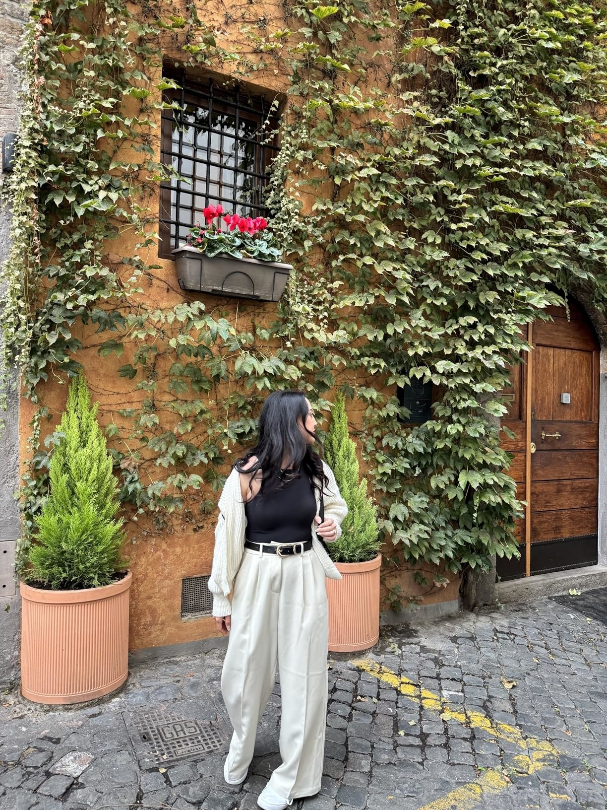 A reviewer wearing a black top and beige pants stands in front of a building with ivy-covered walls and a wooden door, accompanied by potted plants