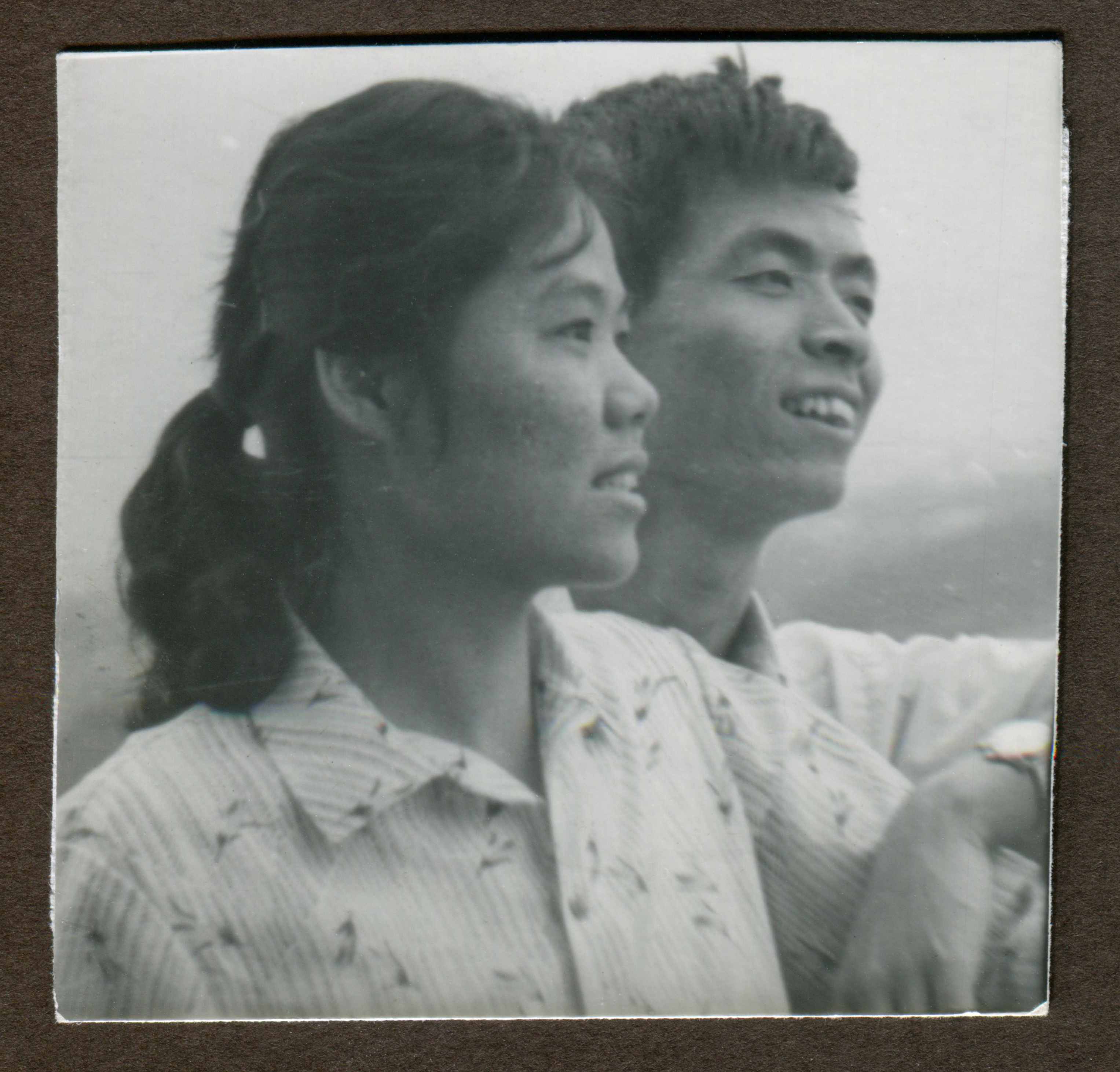 A young man and woman, both smiling, stand close together, with the woman in the foreground. They are looking into the distance