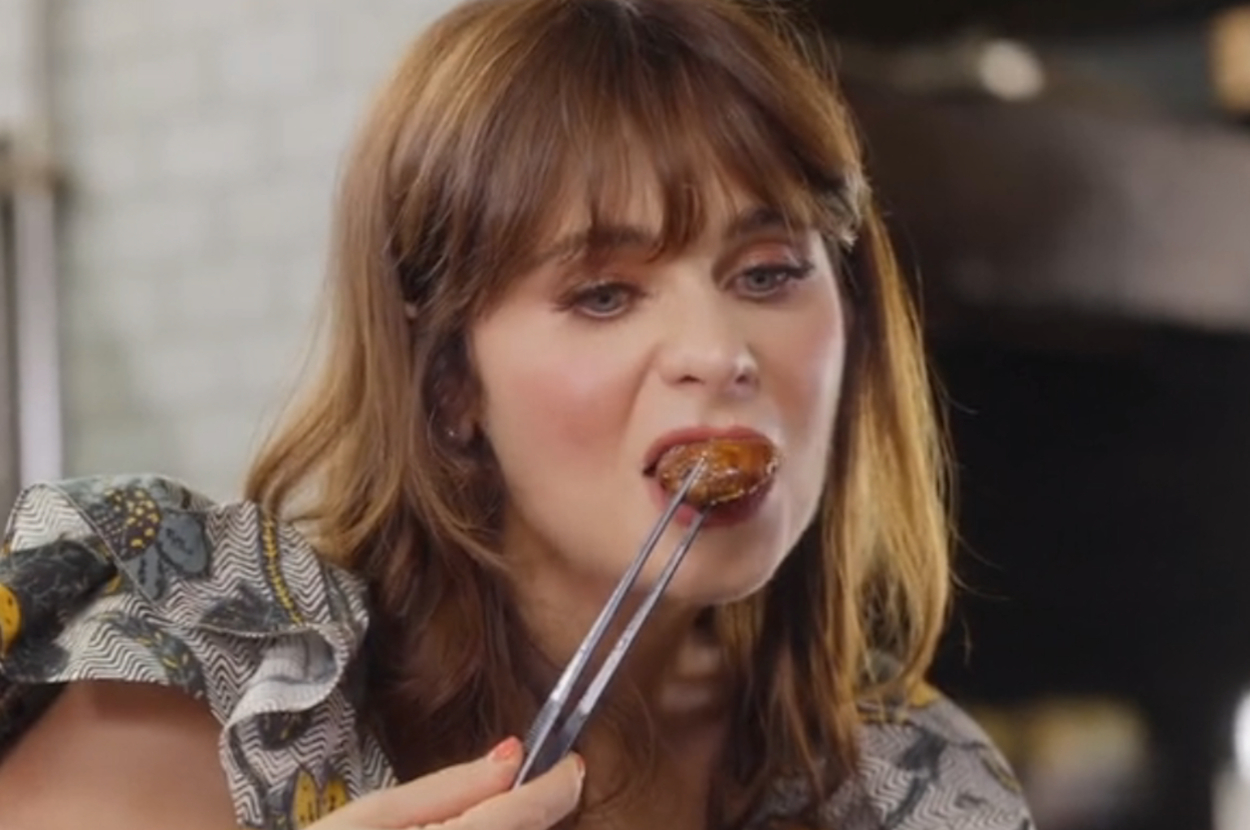 Zooey Deschanel is holding chopsticks and eating a piece of food, looking thoughtful. She has wavy hair and is wearing a patterned blouse with ruffled sleeves