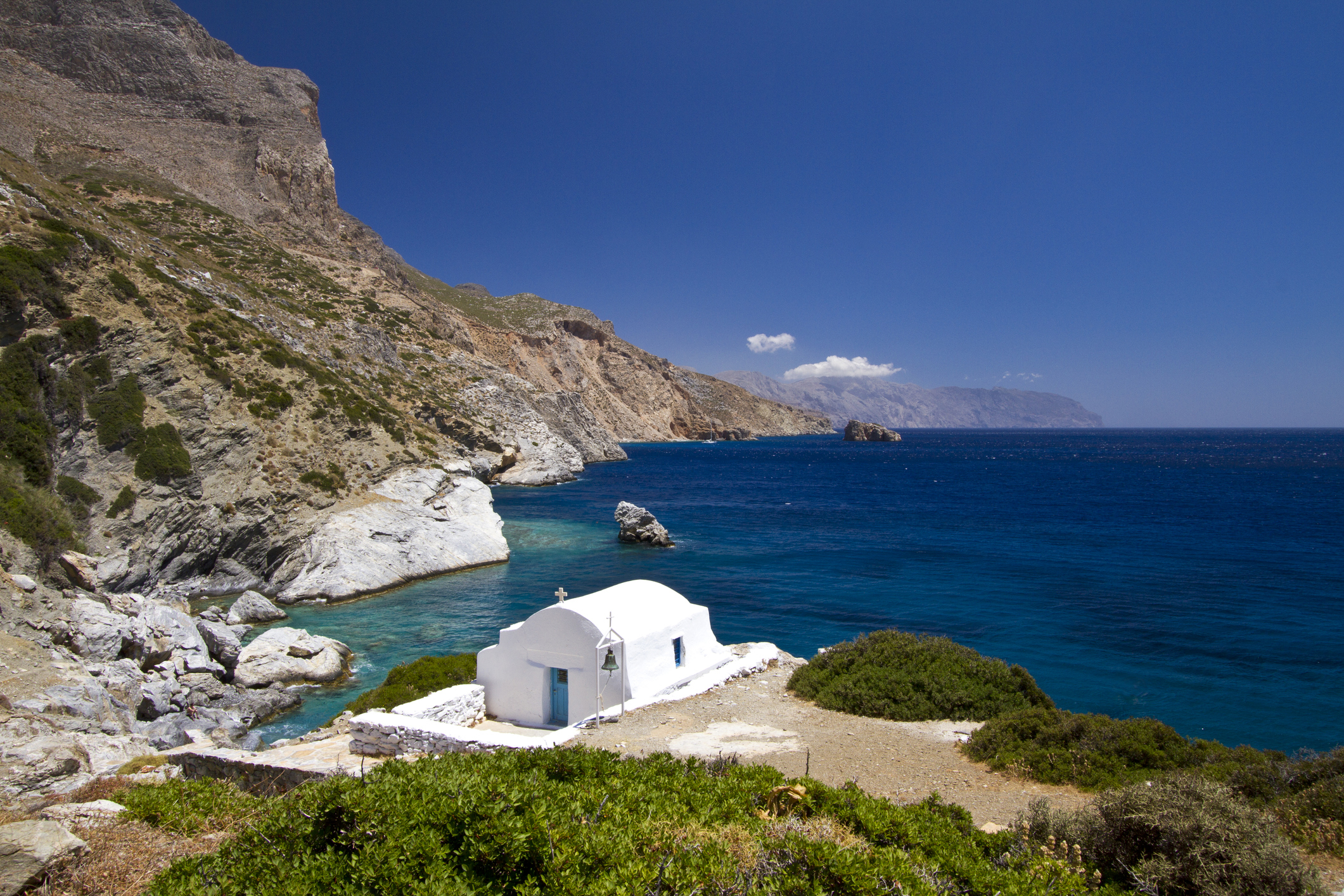A white chapel sits on a rocky coast with a blue sea and mountainous background under a clear sky