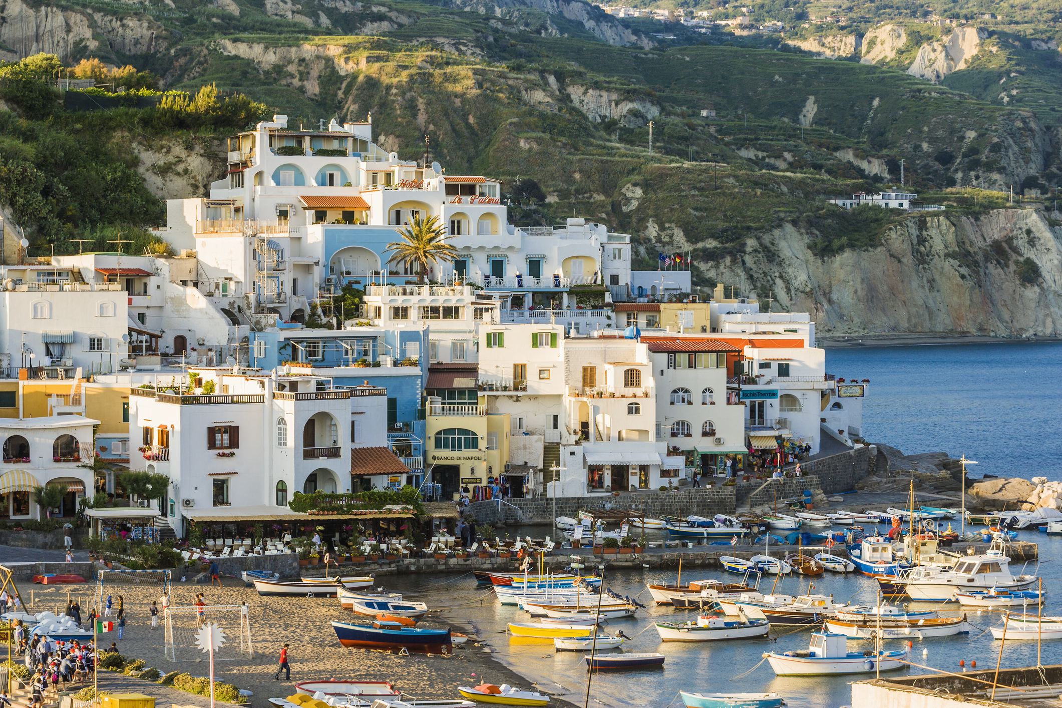 A coastal village with multi-story buildings overlooking a harbor filled with small boats. People are seen walking on the dock. Hills are visible in the background