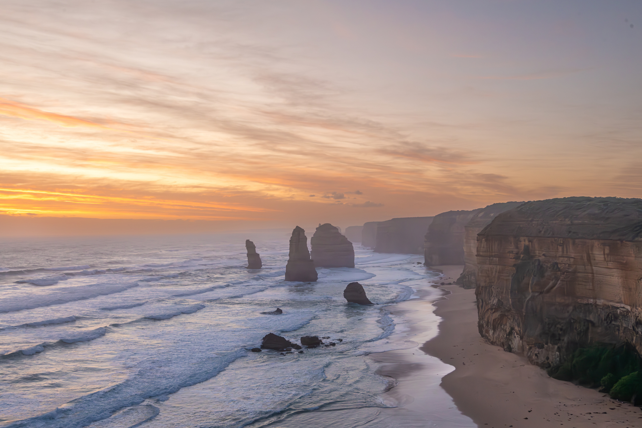 A scenic view of the Twelve Apostles rock formations along a coastal shoreline at sunset, with gentle waves rolling in